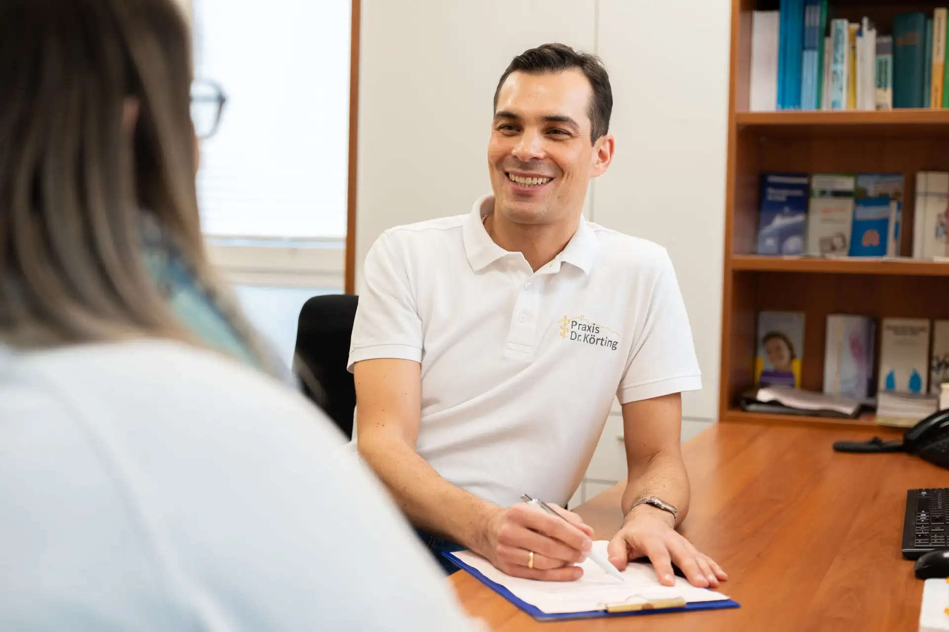 Lächelnder Arzt im Patientengespräch mit einem Klemmbrett auf dem Tisch